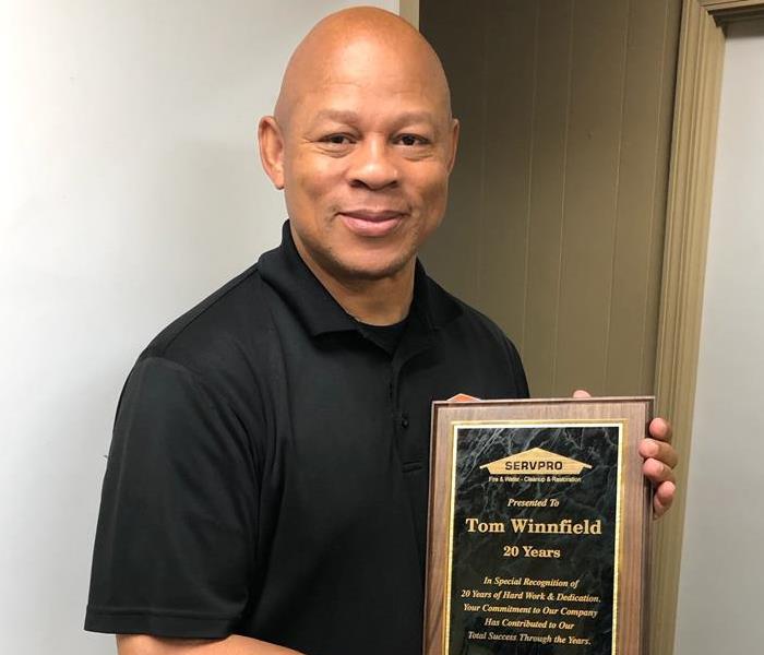 A bald male employee in a black polo shirt holds his 20-year dedication plaque.